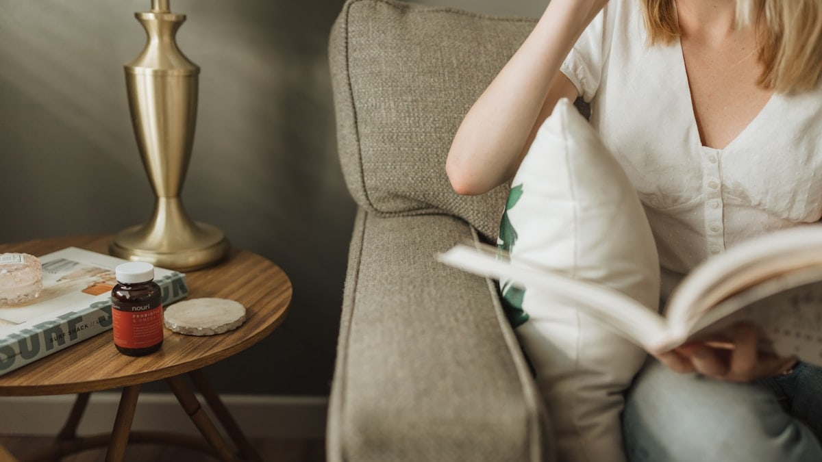 Woman reading book on living room couch in the morning. Side table holds magazines, a lamp, and a Nouri Gut Rest probiotic bottle.