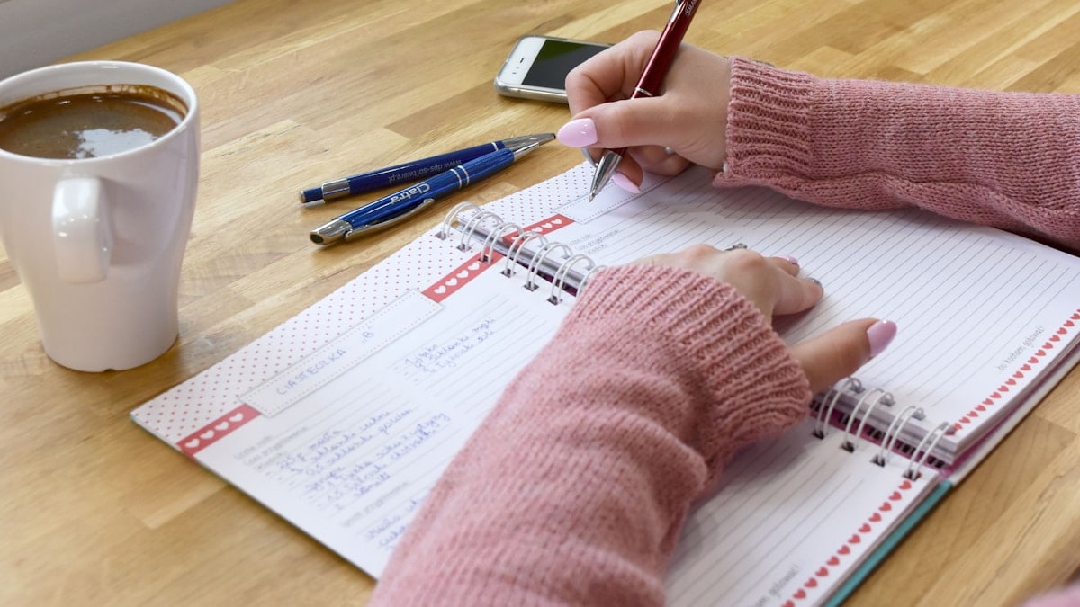 A woman in a cozy pink knitted sweater writing a recipe in a notebook on a wooden table, with a cup of coffee and pens nearby. Cozy morning atmosphere, home office, or meal planning concept.